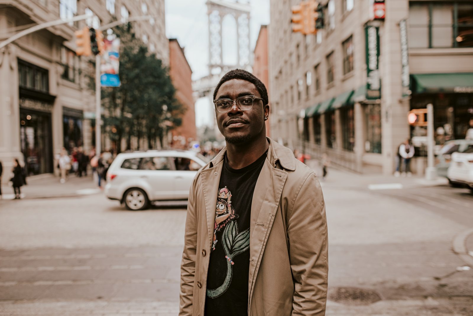 Shawn in a camel trench coat and glasses on a Dumbo street, the Manhattan Bridge behind him