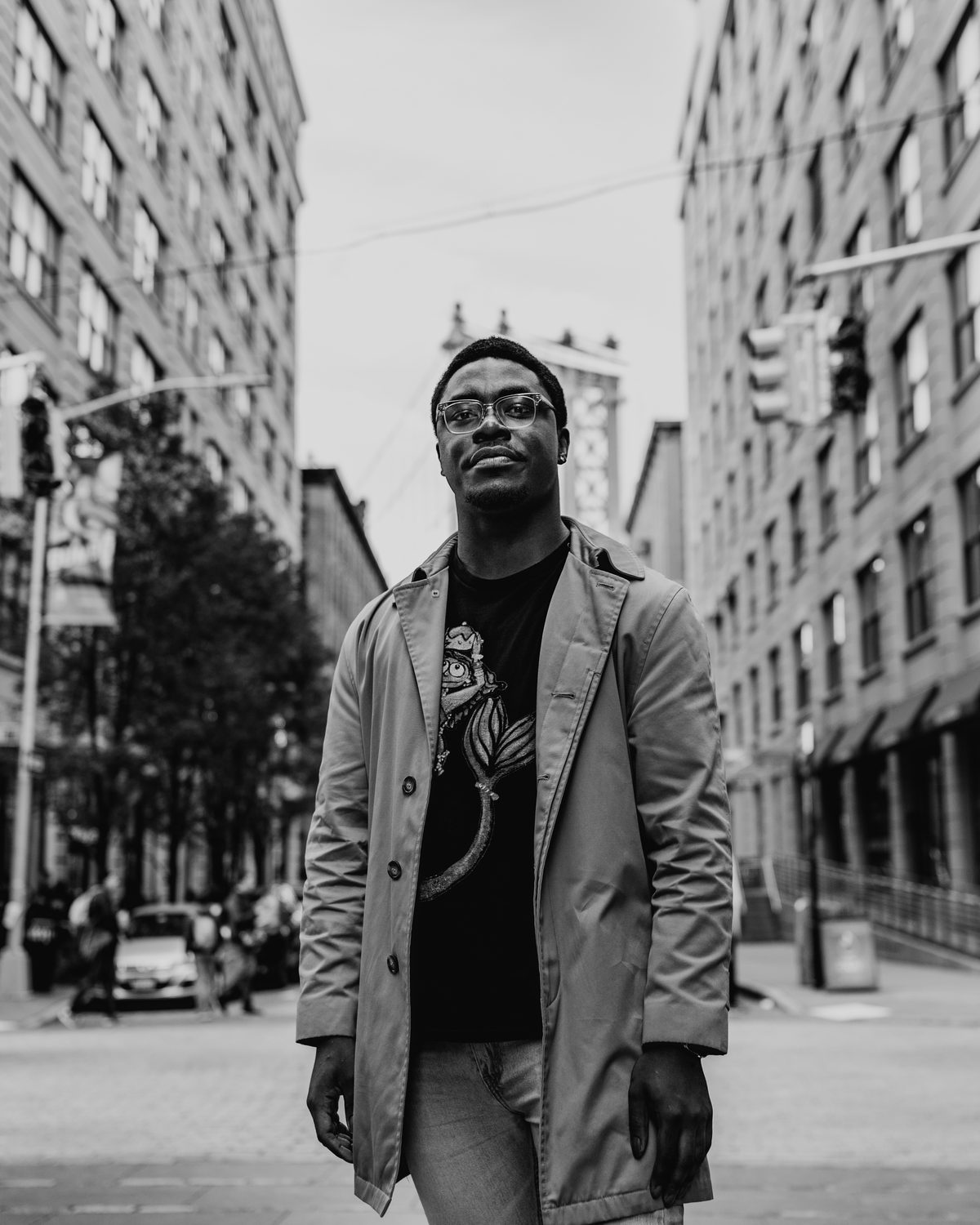 Shawn Smith Jr. in glasses and a trench coat, standing in a Dumbo street with the Manhattan Bridge behind him, in black and white