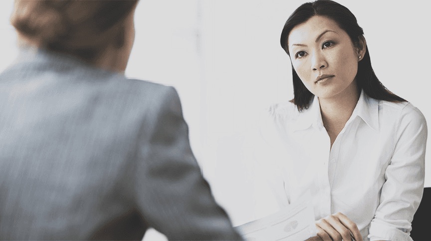 A woman in a white shirt facing an interviewer across a table, unsmiling