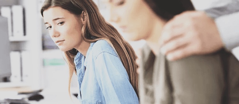 A young woman sitting apart, distressed, while two colleagues comfort each other